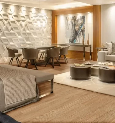 Modern living and dining room with a geometric white wall, glass dining table, gray chairs, beige sofa, round dark ottomans, and abstract art.